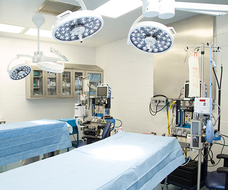 Two operating tables and various surgical equipment in a beige, cinder-block OR