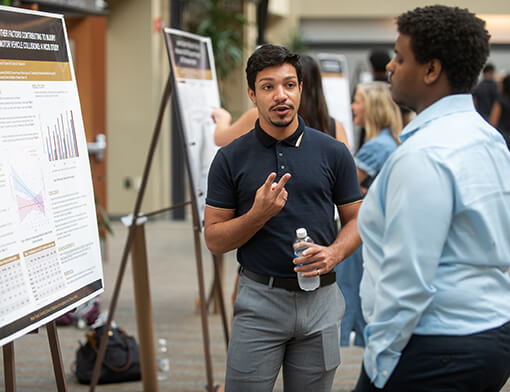 A young man with dark hair wearing a dark gray polo speaks to a man in a light blue shirt about data on a research poster.