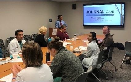 Several people sit around a conference room table with 'Journal Club' projected on a screen behind them