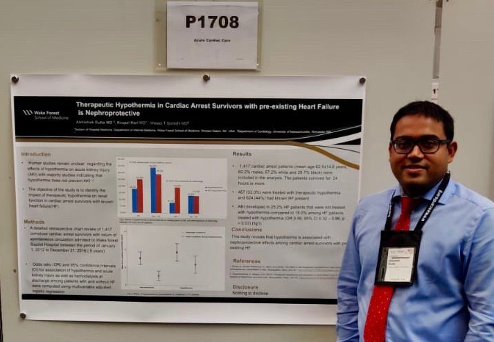 A man in a blue button-down shirt and red tie smiles as he stands in front of a research poster