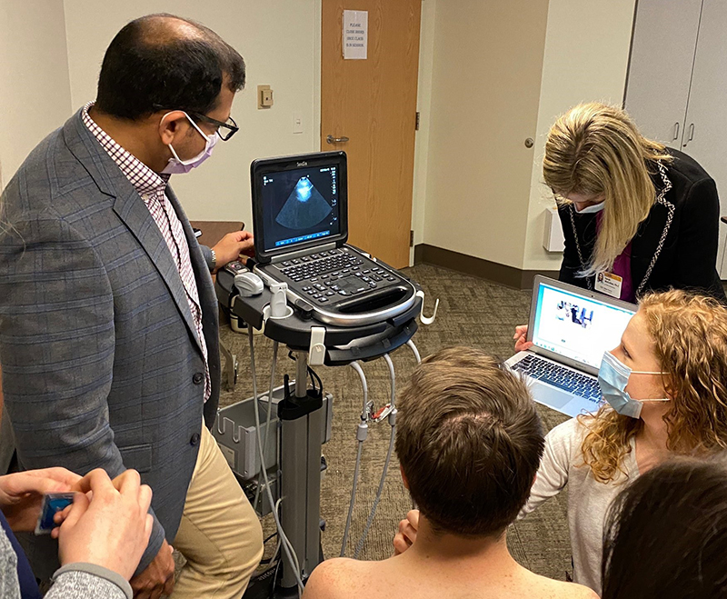 A man looks at an ultrasound machine as a woman leans over a laptop computer display. Younger people, including a shirtless man, sit in a semicircle and look at the ultrasound machine