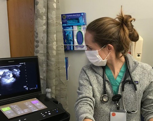 A woman wearing green scrubs, a fleece pullover, a stethoscope and a face mask turns halfway to look at an ultrasound machine