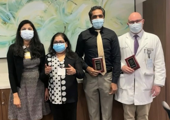 Two men holding plaques and two women (all wearing face masks) stand in front of an abstract painting and pose for a photo