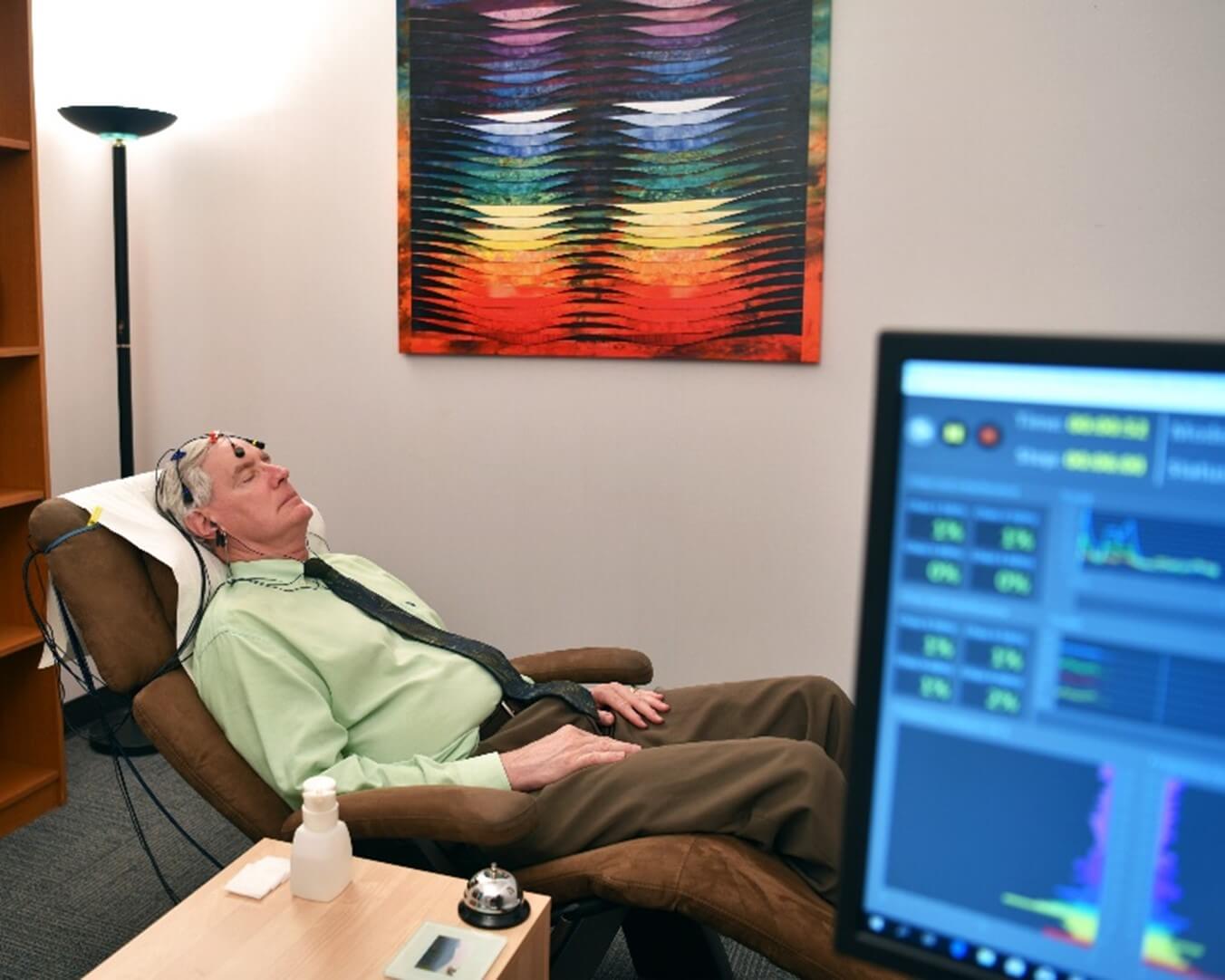 An older man in a green collared shirt lies down on while having monitoring devices attached to his head.