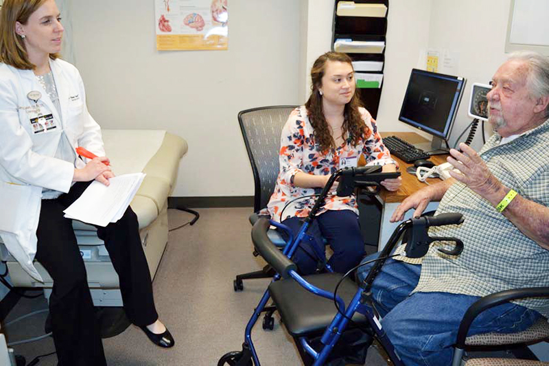 A woman in a white coat and another in a floral top listen to an older man talk in an exam room