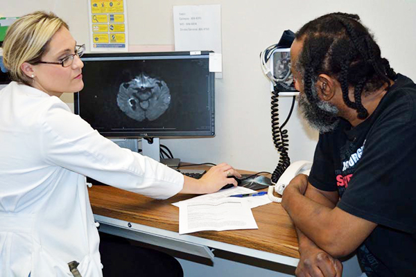 A blonde female in white coat talks to a Black male patient with an MRI image of a brain on a computer screen