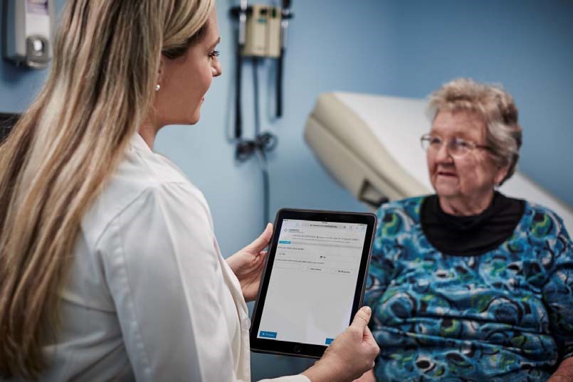 Female doctor wearing white coat and with long blonde hair talks to older woman in a blue exam room