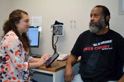 Woman in a floral shirt enters information on a tablet while talking with older Black man in exam room