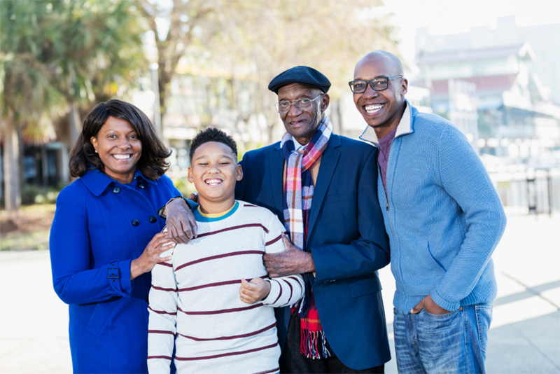 A Black family consisting of a grandfather, mother, father and teenage son, stands together and smiles at the camera. The adults are wearing shades of blue and the teen is wearing a horizontal striped sweater