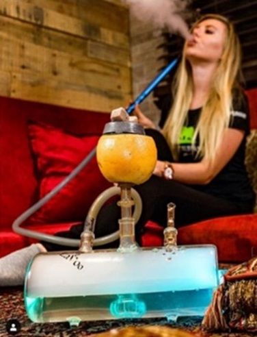 Girl with long blonde hair exhales a plume of smoke in the background and a water tank smoking device sits in the foreground