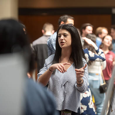 A young woman with dark brown hair talking to a large group of people.
