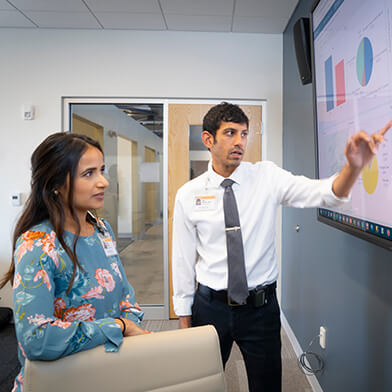 A young woman in business casual attire listening to a lecture given by a young man in a white shirt and dark tie.