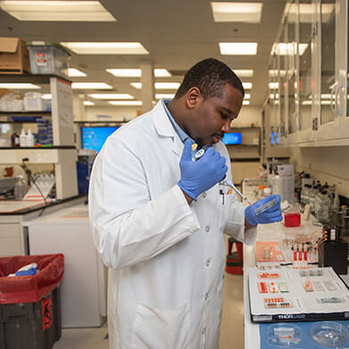 A man wearing a white lab coat and blue gloves examines a collection of samples in a lab.