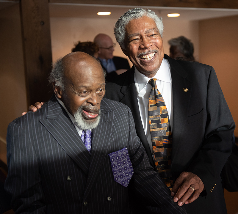 Two African-American men with grey hair stand together and smile 