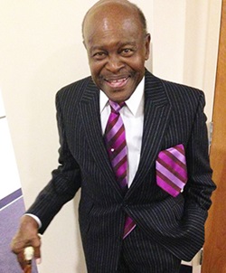 A dapper older African American gentleman wearing a dark suit with matching purple-striped tie and handkerchief smiles at the camera