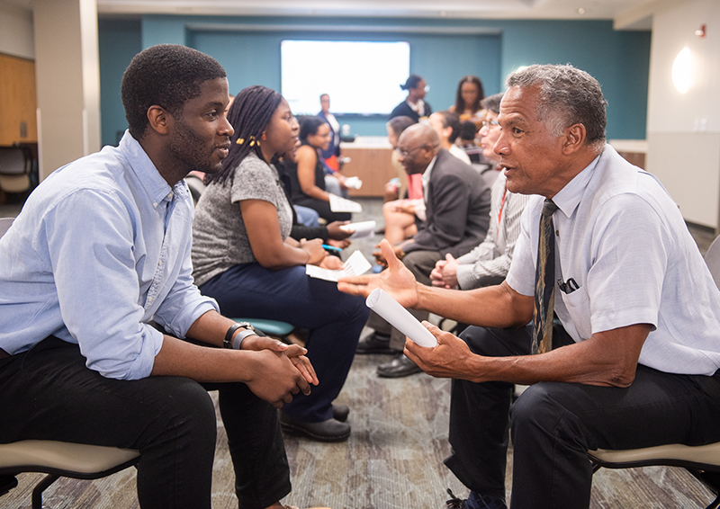 Two African American men, one younger, one older, sit face-to-face with knees almost touching and the older man talks and gestures