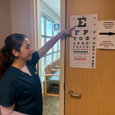 A young woman in dark blue scrubs pointing at the top of a vision testing sign.