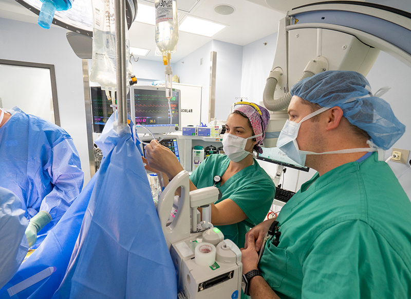 Two people in green scrubs and PPE monitor equipment in the OR