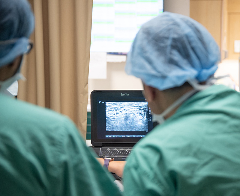 Two people in green surgical scrubs and hair nets look at a monitor