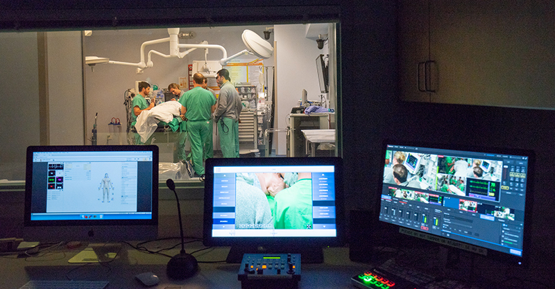 Three computer monitors glow in foreground in dark room and large window looks into simulated operation with multiple people in green scrubs