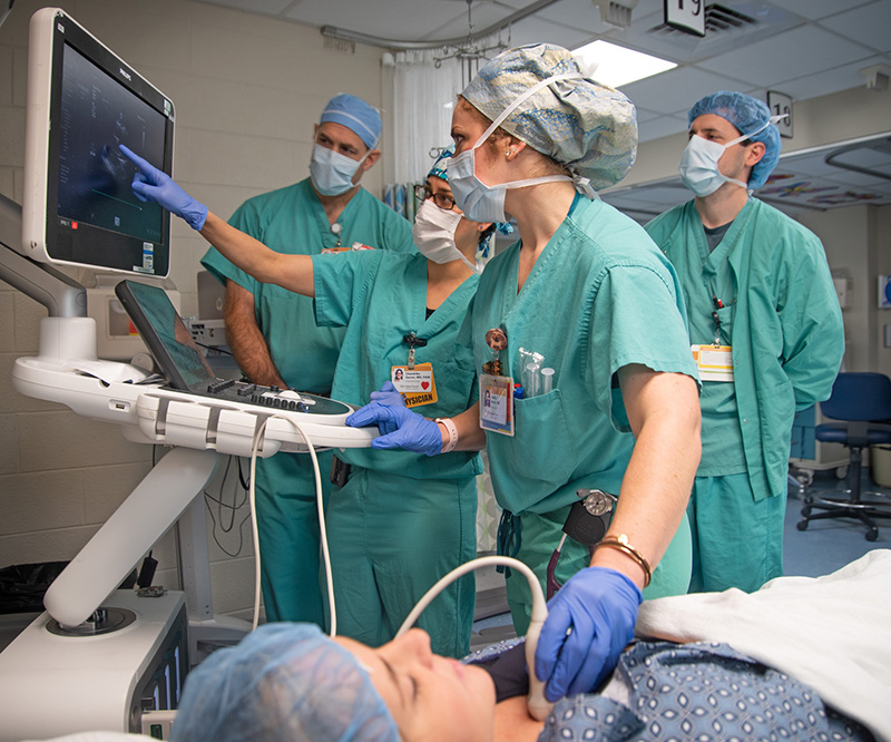 Four people in green scrubs watch a monitor while checking activity on a sleeping patient