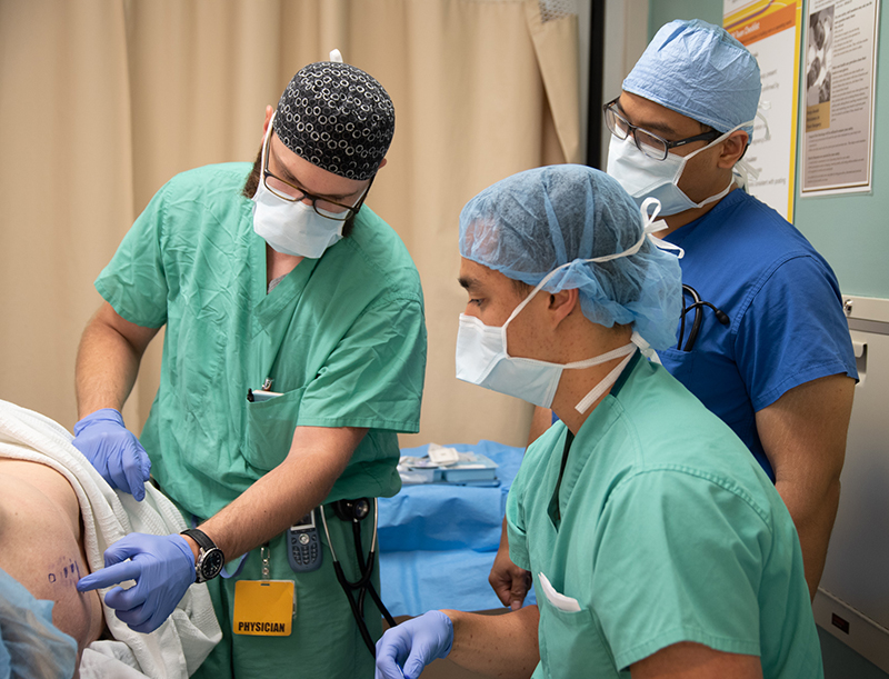 Three people in surgical scrubs examine markings on a patient's spine