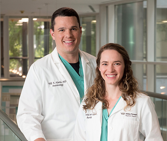 A caucasian man and woman in lab coats smiling at the camera in a window filled hallway.