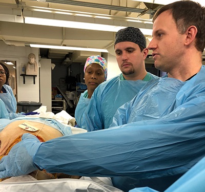 Several people in blue scrubs stand around and look at a cadaver