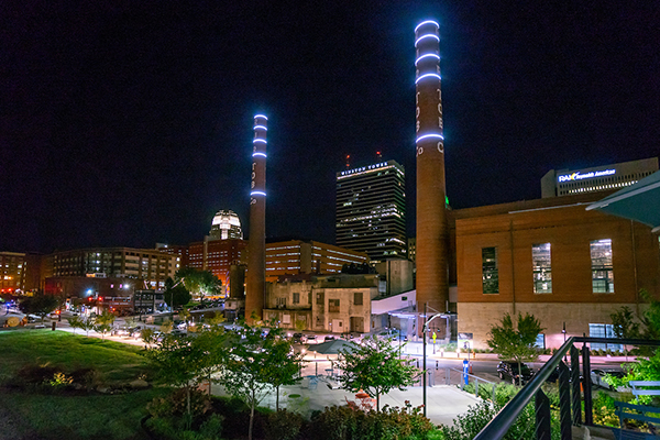 Two brick smokestacks and an industrial brick building are lit by LED lights against a nighttime skyline of Winston Salem