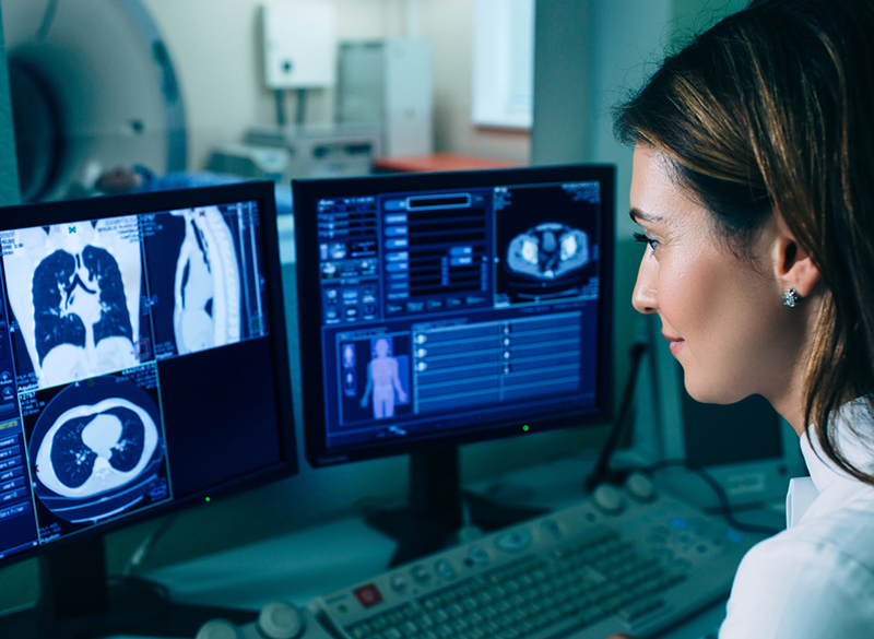 Dark-haired woman looks at blue-gray images from a CT scan on a computer screen, and blurred in the background is CT machine in another room