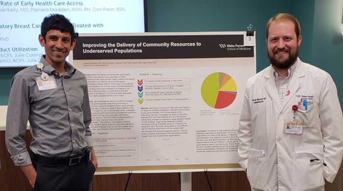 Ajay Dharod, MD, and Andrew Benefield, MD, stand in front of a research poster for 'Improving Social Determinants of Health through the Delivery of Community Resources' with a projection screen behind them.