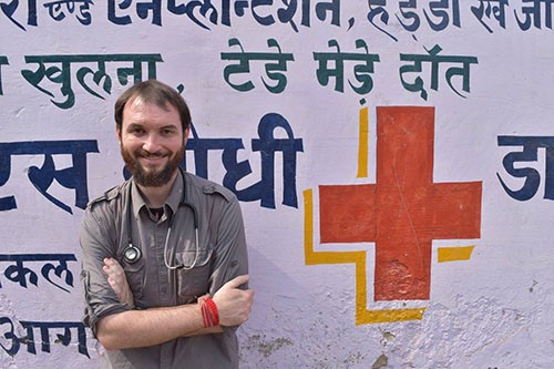 Bearded white man smiles and stands in front of wall with Red Cross symbol and Arabic writing