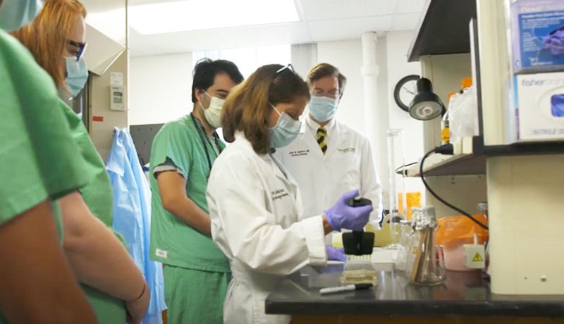 A woman wearing a white coat, gloves and face mask handles a pipette in a lab as people wearing green scrubs and face masks stand around her