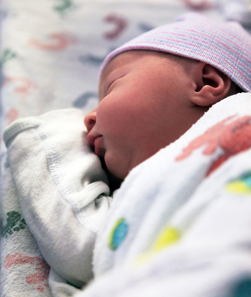 Newborn wearing longsleeve onesie and cap sleeps in isolette