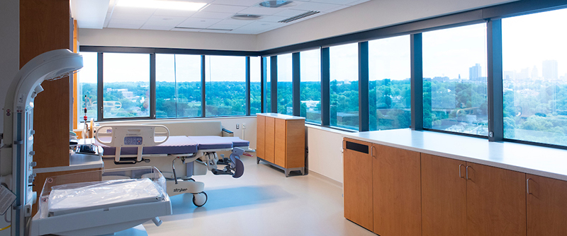 Large corner room with windows on two walls, a labor and delivery bed and a warming bassinette