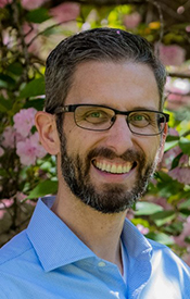 Bearded Caucasian man with glasses stands outside and smiles