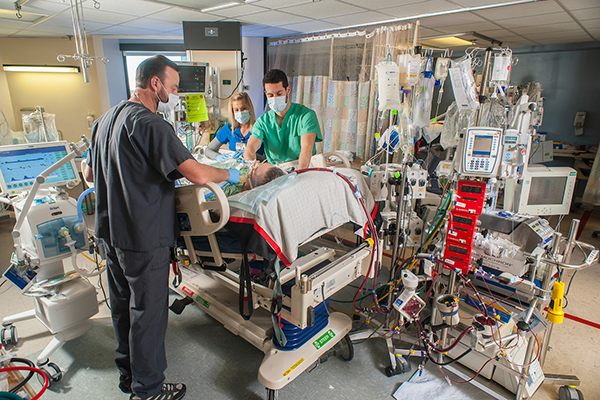 Two men and a woman in scrubs and masks stand around a patient in an ICU bed, surrounded by medical equipment