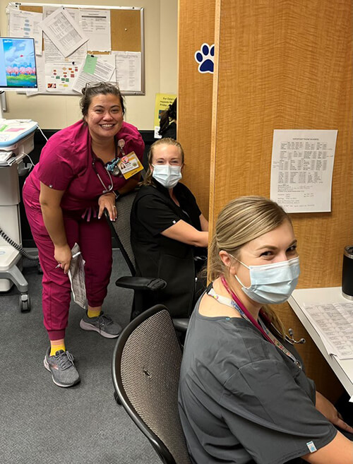Three medical professionals wearing scrubs and sitting at a desk looking at the camera..