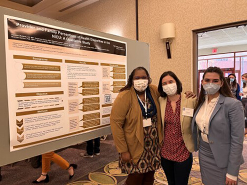 Three women wearing masks, standing side-by-side in front of a presentation.