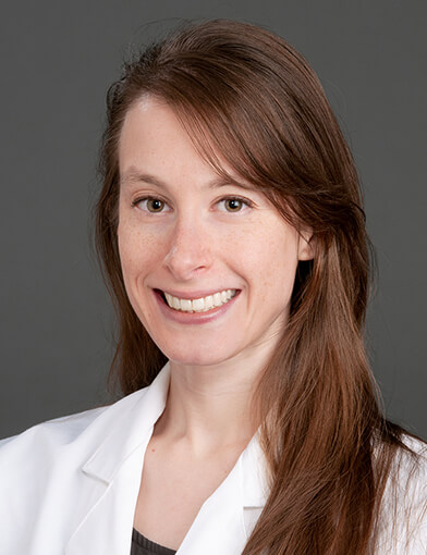 A woman with long hair, wearing a lab coat and smiling at the camera.
