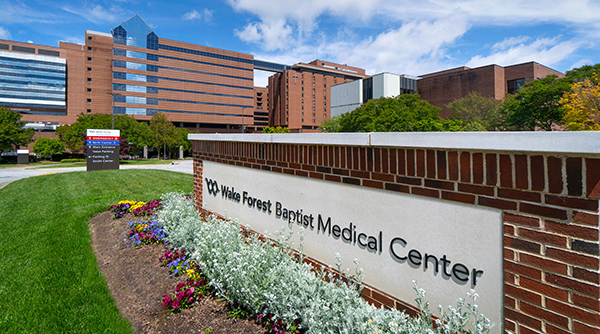 brick buildings sit in the background and a 'Wake Forest Baptist Medical Center' entrance sign is in the foreground and surrounded by flowers