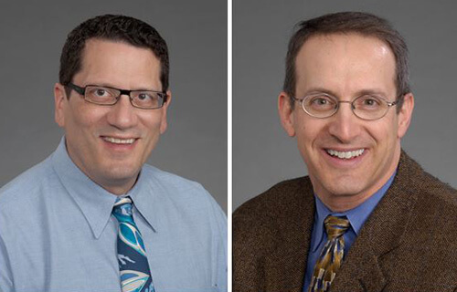 Two older men both wearing glasses, sitting and smiling at the camera. One is wearing a blue shirt with a tie and one is wearing a brown jacket.