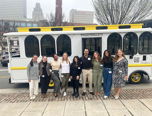 Medical students who attended the Second Look event stand for a group photo in front of a white, blue, and yellow trolley.