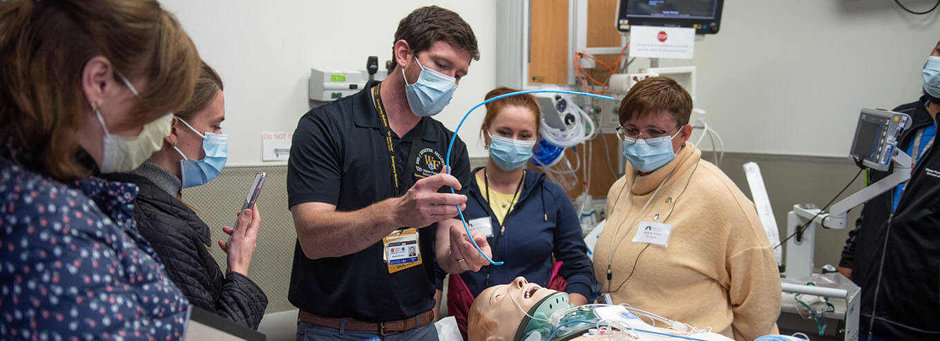 Medical students and faculty in masks performing procedures on a test dummy in a classroom setting.