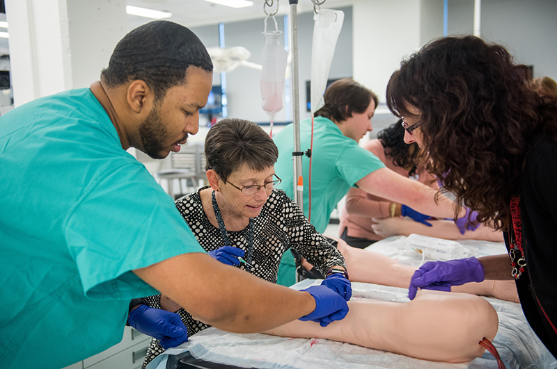 A Black man in green scrubs and gloves demonstrates something on an artificial leg for two women in the anatomy simulation lab