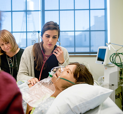 A woman wearing a stethoscope listens to a mannikin at a cardiopulmonary simulation event