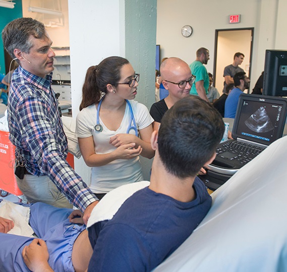 A group of medical students look at an ultrasound monitor as an instructor uses an ultrasound wand on a volunteer