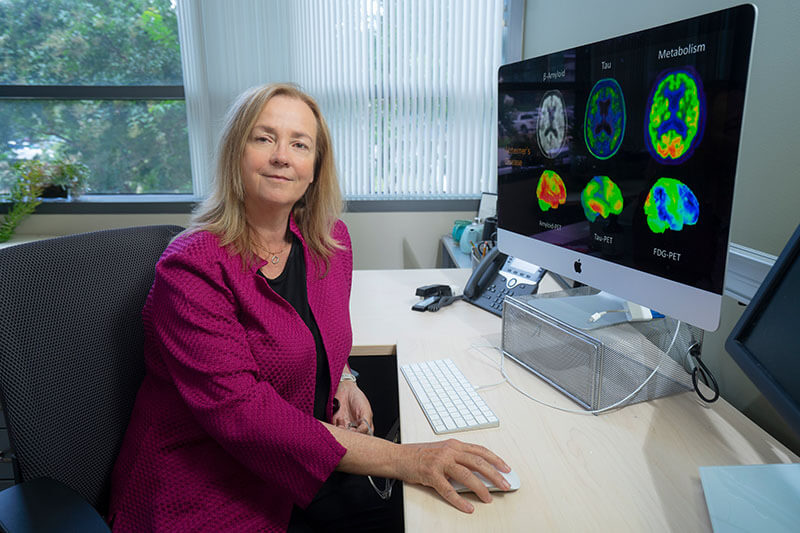 A woman in a pink shirt looking at the camera sitting next to a computer with brain scans on the screen.