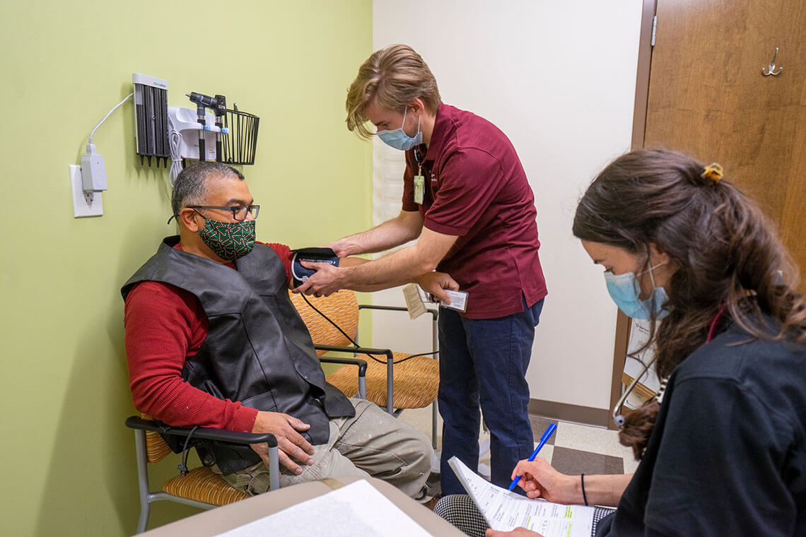 A patient with a gray vest and red long sleeve shirt getting his blood pressure checked by a nurse in a red shirt and blue pants while another staff member observes.