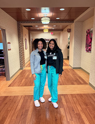 Two women wearing dark blue shirts and light blue scrubs standing together in a hallway.